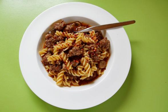 Ein tiefer Teller mit Nudeln, Pilzen, Fleischbällchen und Brühe. Der Teller steht auf einer grünen Tischplatte. Ein Löffel steckt in den Nudeln.