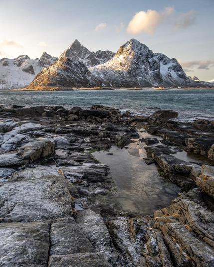 The image shows a stunning landscape featuring snow-capped mountains, likely part of a rugged mountain range near a body of water. In the foreground, there's a rocky shoreline with a small pool of water. The lighting suggests it may be taken during the golden hour, either sunrise or sunset, as parts of the mountains are warmly lit. The scene conveys a sense of calm and natural beauty.