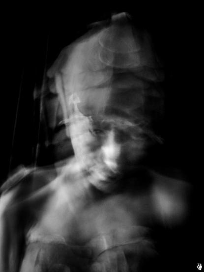 bw photo, dark background. headshot in portrait format of a woman, with headgear made of leafy scale-like parts, blurred by motion and looking slightly from below upwards at the viewer. Her gaze is calm but somewhat unsettling.