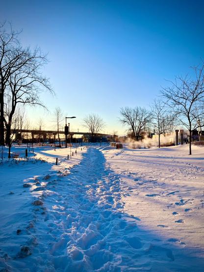 A snow-covered path winds through a winter landscape, flanked by bare trees against a clear blue sky. The sunlight casts a warm glow on the snow, creating a serene and tranquil atmosphere. In the background, steam rises from a nearby structure, adding to the chilly yet picturesque scene.