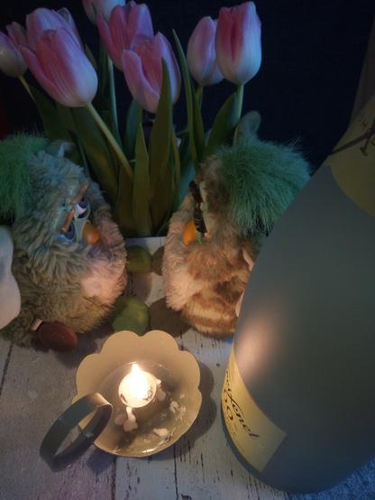 Zwei grüne Furbys sitzen einander gegenüber. Hinter ihnen sind weiß-lilane Tulpen. Vor ihnen steht eine brennende Kerze in einem grünen Kerzenhalter. Rechts vor den Furbys steht eine Buddel alkfreier Sekt.