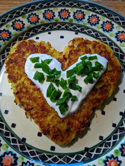 Na kolorowym talerzu z kwiecistą obwódką leży, wysmażony na rumiano, placek ziemniaczany w kształcie serca, a na nim biały kleks z jogurtu naturalnego posypany siekanym grubo zielonym szczypiorem.