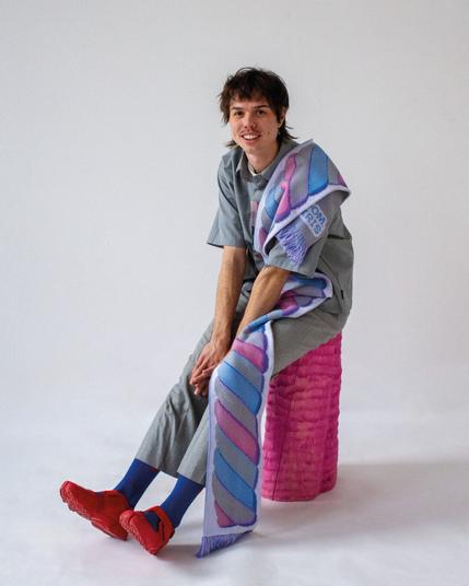 a studio photo featuring a young man wearing a scarf that has a 'spekje' a dutch sweet snack that tastes like a marshmellow on it. the model in the photo is Chris Tam Nelck, he's sitting on a pink piece of wood & is wearing bright red tabi-shoes.