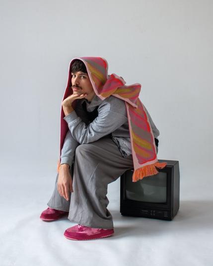 another studio photo of a young man wearing the same 'spekje' scarf, this time in a pink/orange color. he's sitting on an old LCD tv, he's wearing pink shoes. the model in the photo is Robin Rosbak, he's also the stylist of the shoot.