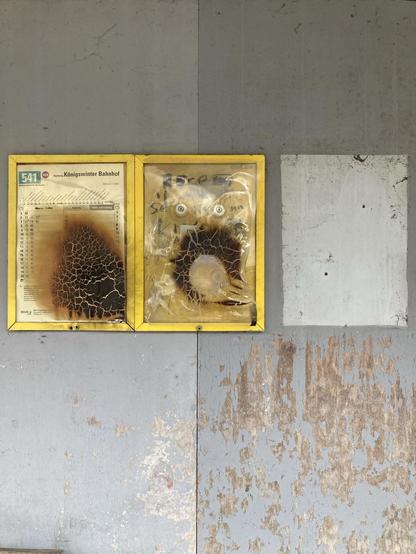 Fotografie der Busfahrplanrahmen im Inneren einer alten Bushaltestelle. Der Hintergrund ist grau und teilweise abgeschabt, dass die darunter liegende Holzplatte zu sehen ist. Links und mittig hängen zwei gelbe Rahmen mit verkohltem Plastikschutz. Die Busabfahrtszeiten  sind am Rand gerade noch lesbar. Rechts daneben ist ein helles graues Feld zu sehen, in Rahmengrösse, wo früher einmal ein Rahmen gehangen haben wird.