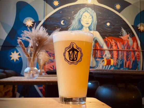 A glass of L’Amoureux #3, a New England DIPA, with in the background a wall painting done by INDEXPOUCE, picturing in pink and blue, a naked lady emerging from a pond, pouring beer into it from a glass with handle in each hand.
