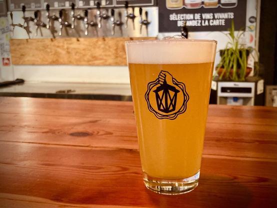 A glass of L’Amoureux #1, a New England pale ale, with in the background the taps of the L’Ermitage taproom.