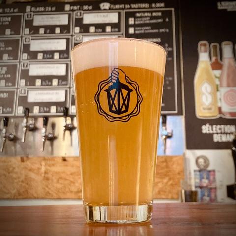 A glass of L’Amoureux #2, a New England IPA, with in the background the beer board behind the bar of the L’Ermitage taproom.