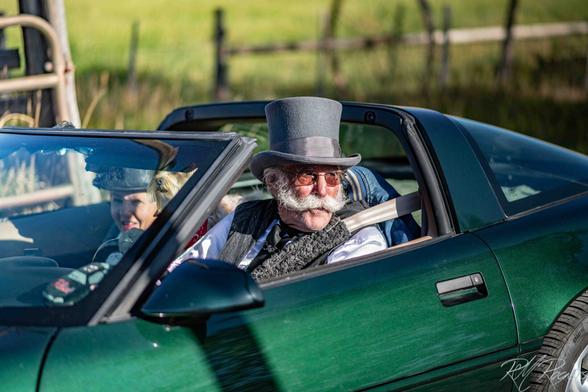 An older couple in a green corvette are dressed in fanciful clothing.