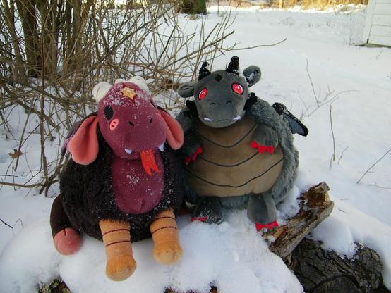 Two plushies, one reddish pink and brown with orange feet and white horns, the other one a grey dragon with red eyes.
