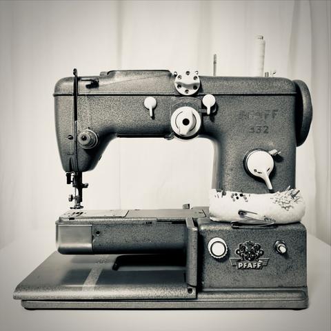 Schwarz-weiß Fotografie einer alten Pfaff-Nähmaschine mit Metallgehäuse in Hammerschlagoptik. Es ist weißes Garn eingespannt und ein kleines mit Nadeln gespicktes Kissen rechts am Körper der Maschine befestigt.