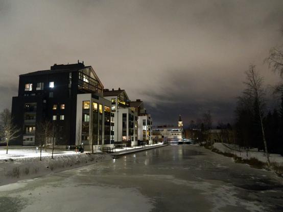 A view of a frozen canal at night time in winter, with heavy overcast brown sky. An apartment building is on the left side of the canal, with barren trees on the right side. In distance, the city centre with church clock tower can be seen.