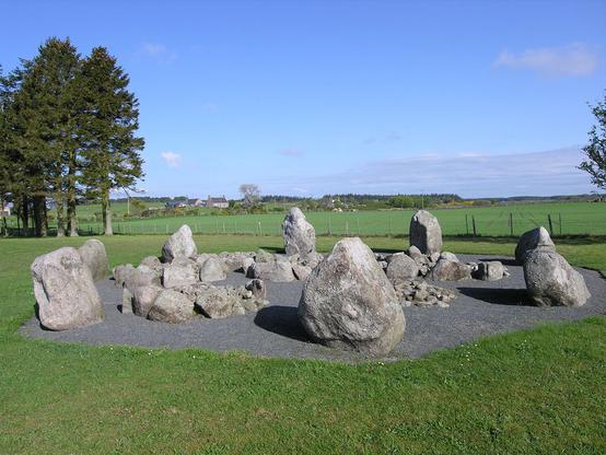The Cullerlie Stone Circle near Echt. The image shows a grassy area containing a gravel octogon, within which is a circle of low standing stones, and within that are many smaller stones. There are some trees to the left rear and the sky is nearly clear blue.