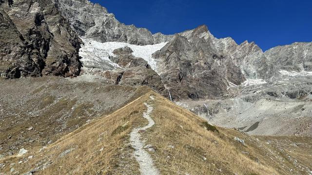A narrow footpath leads along a sharp-crested ride toward a mountain wall with a small white glacier.