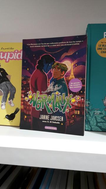A photography of a graphic novel on a bookshop shelf. It's intitled "Les Normaux" in green with littles stars around it, and it features two characters (a blue skin and dark haired vampire and a green haired human) standing in front of each other with Paris in the background.