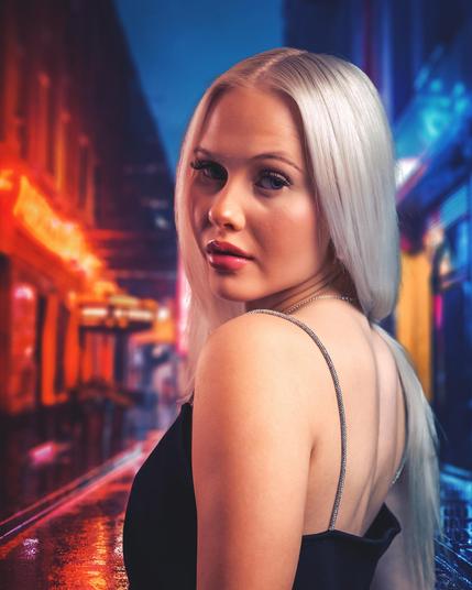 The image shows a person with long, light-colored hair, wearing a black outfit. The background appears to be a vibrant, neon-lit street, giving the scene a night-time city vibe. The colors are predominantly blues and reds, contributing to a moody, urban atmosphere.