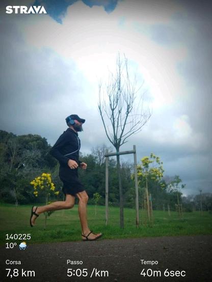 Un runner in sandali e tutto vestito di nero corre in un parco con sullo sfondo degli alberelli di mimose in fiore e un orizzonte imbiancato da grandi bassi nuvole.