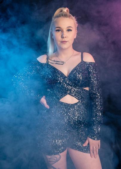 The image shows a person posing in a sparkly, black outfit. The scene is set with colorful, atmospheric smoke in shades of blue and purple. The person has long, light-colored hair tied up and there is a tattoo visible on their chest.