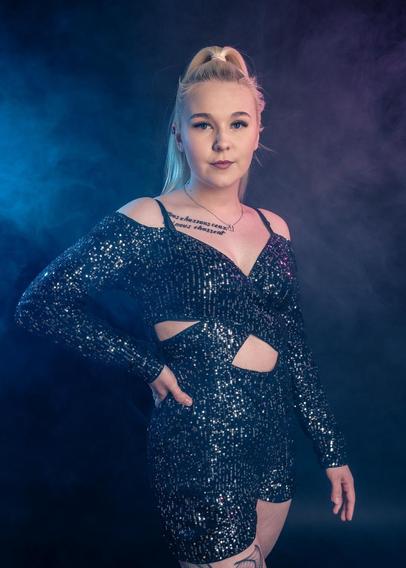 The image shows a person wearing a sparkly outfit with cut-out details, posed in front of a moody, smoky background with blue and purple lighting. They have blonde hair styled in a high ponytail.