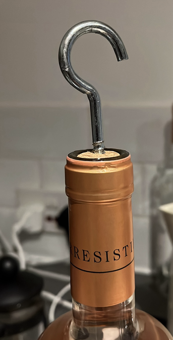 A closeup of a wine bottle with a cork, there is a hook screw stuck on it. The neck of the bottle has a golden label and you can see the text "resist" in black letters. You can see a white kettle and a tiled wall in the background.