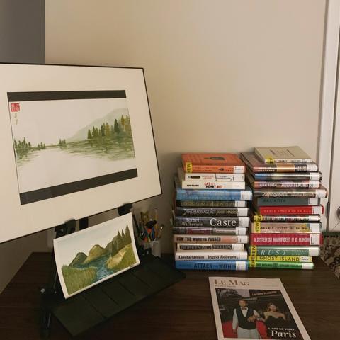 Two pieces of art displayed on an easel on the left side of a dark wooden desk and two stacks of library books and a magazine on the right side of the desk. All against a blank white wall