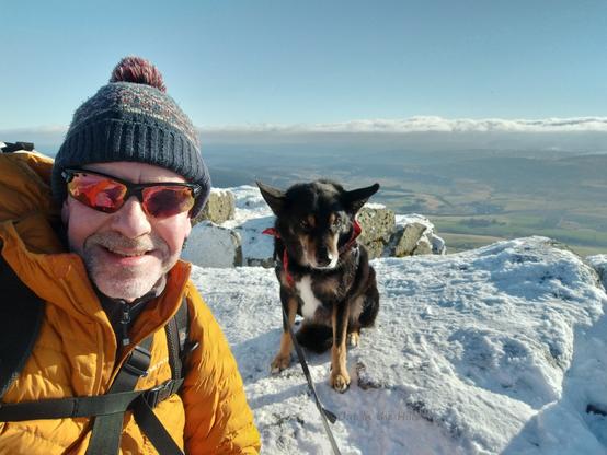 Summit selfie, Moray the dog and me. We are sat on rocky ground which is covered in snow. I am wearing sunglasses and wrapped up well against the cold wind, with yellow padded jacket and woollen bobble hat. In the background the lower ground is hazy but clear of snow. Hazy blue sky with clouds onthe horizon.