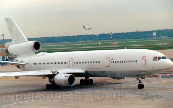 Side view of a white, 3 engined jet airliner with a silver belly taxiing from left to right.
The plane is almost entirely blank, only red outlines on the doors, the registration "F-BTDB" on the rear fuselage, and a small red "EAL" logo in front of the mid-forward door providing a break from the "polar bear" livery.
In the background, green grass leads off to trees in the distance under a grey sky, with a white, twin-engined jet airliner with a blue tail on final approach to a runway visible in the mid-distance.