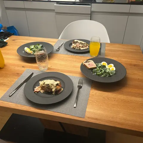 A photograph of a wooden dining table set for three people with black plates and grey placemats. Each plate contains a meal: the main dish appears to be a piece of meat or fish covered in a creamy sauce, with bacon on the side. There are also side dishes of fresh spinach salad with sliced boiled eggs and a slice of pâté or terrine on toast. The table has beverages, including a glass of water and a glass of orange-colored juice.