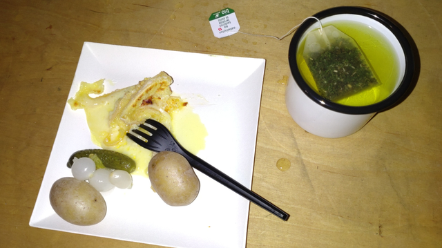 Raclette mit kleinen Kartoffeln und Mixed Pickels (Essiggurke und Silberzwiebel) auf einem quadratischen Pappteller und mit schwarzer Plastikgabel sowie Salbeitee in einem weissen Metallbecher mit schwarz lackiertem Rand.