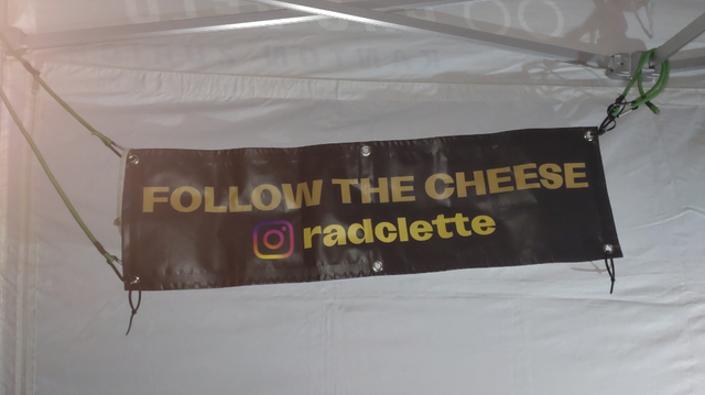 Ein Banner mit der Aufschrift "FOLLOW THE CHEESE [Instagram Logo]radclette" in einem weißen Partyzelt mit Rückwand.