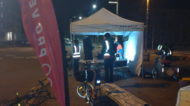 Ein beleuchtetes, weisses Partyzelt im Dunkeln mit diversen Menschen in zum Teil reflektierender Kleidung. Im Vordergrund ein halb gefaltetes Brompton-Faltrad mit reflektierender Spirale an der Lenksäule und Satteltasche mit reflektierendem Aufnäher, ein liegendes Velo und eine Pro Velo Strandflagge. Im Hintergrund ein Lastenrad mit einer Person mit reflektierender Kleidung und reflektierendem Velohelm.