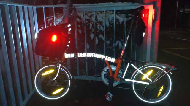 Ein orangefarbenes Brompton steht an ein Metallgeländer mit Schneekrönchen gelehnt. Zwei rote (am Sattel und der Fronttasche hinten) und eine weisse LED-Leuchte (an der Fronttasche vorne) sind sichtbar und an. Auf dem Hauptrahmerohr stwht in reflektierenden Lettern "WINTERBROMPTON". Ebenfalls reflektierend sind die Vorderradgabel, der Hinterbau, eine Spirale um die Lenkstange, die Reifen und die gelben Reflektoren in den Speichen. Im Hintergrund ein schneebedecktes Gebüsch.