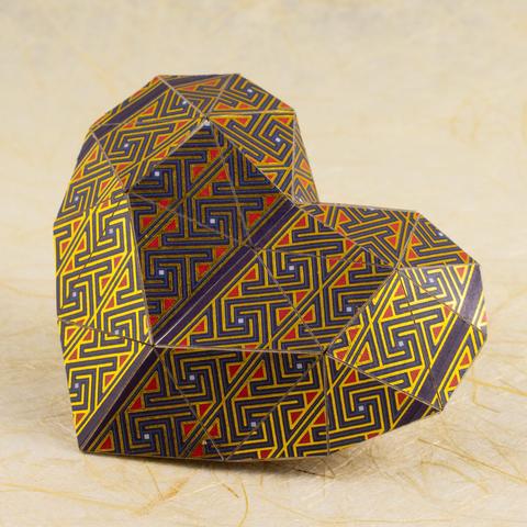 A pepakura (“low poly”) 3D paper heart covered in Celtic key/maze patterns in shades of purple, red, blue, and metallic gold rests on a cream-colored handmade paper background.