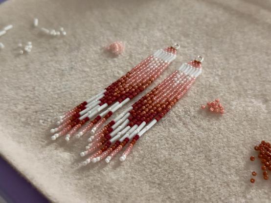 A pair of beaded fringe earrings on a bead mat in white, peach, and burgundy red