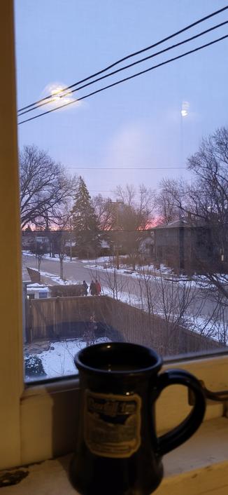 A blue coffee mug sitting on a window sill. Outside is an urban neighborhood with snow on the ground. Three people in winter coats can be seen walking away from the camera toward the sunrise.