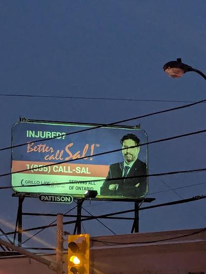 A billboard displaying the text "INJURED? Better Call Sal!", along with a phone number and an image of a man in a suit and tie. At the bottom, the name of the law firm, "Grillo Law," is visible, accompanied by the phrase "Serving all of Ontario." The billboard is mounted on a metal structure, with a twilight sky in the background. In the foreground, a traffic light showing a yellow signal and electrical wires stretching across the image can be seen.