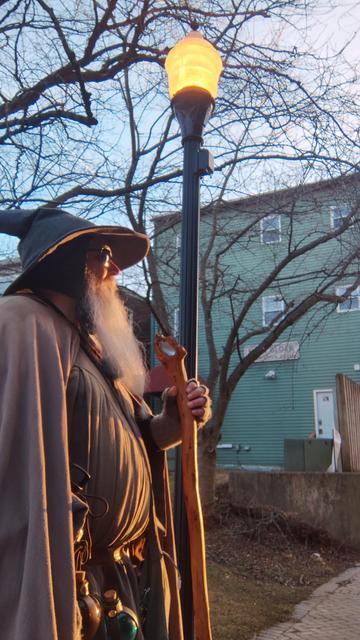 Middle aged man with long grey beard wearing sunglasses and a Gandalf-style wizard outfit with hat & staff looks into evening sunset during golden hour in front of a lit electric street lamp in an alley