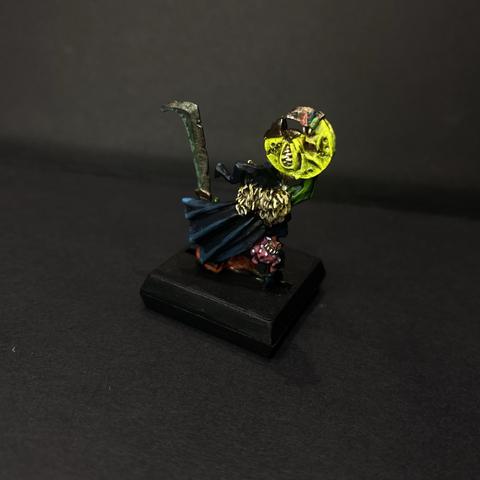Rear of a goblin Mini. There is a black cloak trimmed in fur dominating the perspective, raised above it is a bright yellow shield in the shape of a leering moon. There is also a spotted bag squig on the goblins waist belt.