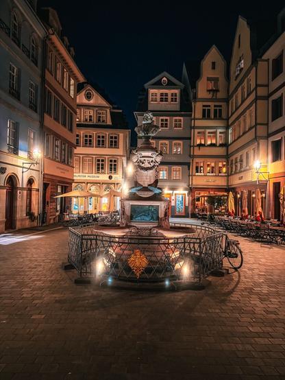 Stimmungsvoll beleuchteter Friedrich-Stoltze-Brunnen auf dem Hühnermarkt in der Frankfurter Altstadt bei Nacht. Der Brunnen mit der Büste des Dichters Friedrich Stoltze steht im Zentrum historischer Fachwerkhäuser, umgeben von Laternenlicht, Restaurants und Cafés. Die warmen Licht-reflexionen und die detailreiche Architektur verleihen der Szene eine atmosphärische Tiefe.