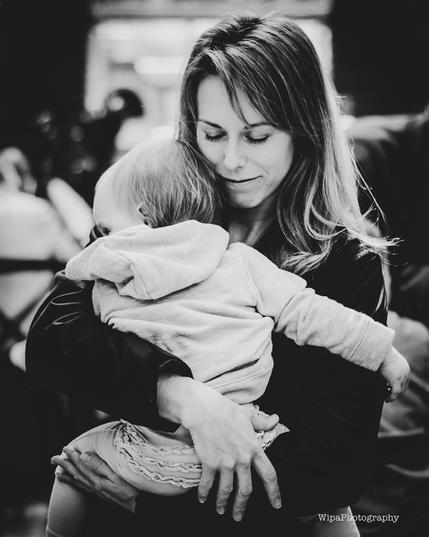 Image of a mother holding a child. She is a toddler and her mom is embracing her with love with her eyes closed. Mom has long hair. Day time.