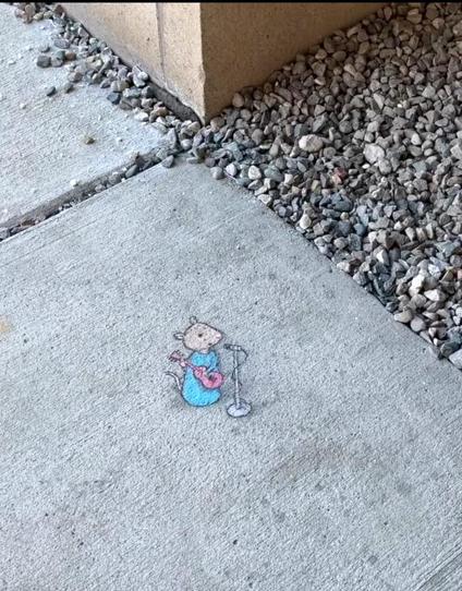 Streetart.  On a gray sidewalk next to little gray pebbles, a little mouse (named Nadine) has been painted with chalk. Nadine stands in front of a microphone in a long blue dress and a red guitar (given her size, it's probably more of a ukulele) and plays music in the direction of the little "rocks". Title: "Nadine's Rock Concert"