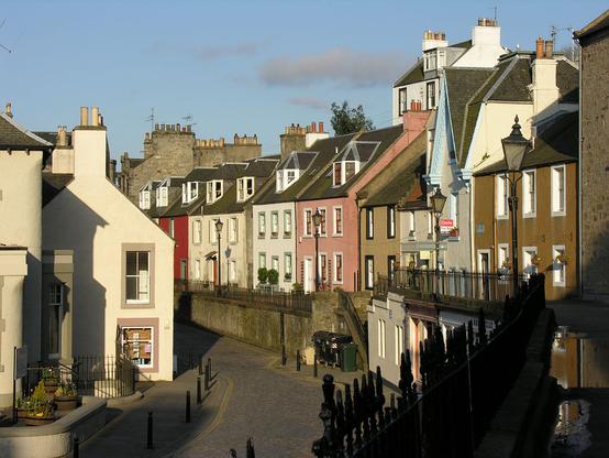 Queensferry High Street. The view is looking along the high street in early morning sunlight. The houses on the right are raised on a terrace above the road and are in browns, pinks and reds as well as white. There is another building on the left, beyond the road and level with it.