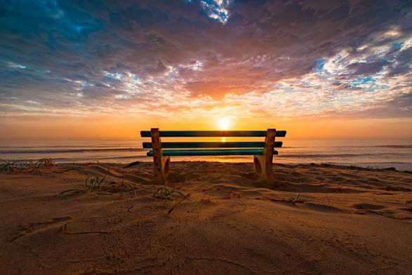 View of a beach at Shoalhaven Heads, Australia, during a summer morning.  A small, teal-colored park bench sits on the sandy beach, positioned near the shoreline.  Low-lying grasses and small shrubs are visible in the sand.  The ocean is calm, with gentle waves.  The sky is a mix of orange, pink, and purple hues, with scattered clouds.  The sun is rising, casting a warm glow over the scene.

The light is soft and diffused, creating a gentle ambiance.  The overall impression is of a tranquil, early morning scene.

Camera: Canon EOS 7D Mark II
Lens: EF-S10-22mm f/3.5-4.5 USM
Settings: ¹⁄₂₅₀ sec at ƒ / 8.0, f/ƒ / 8.0, 10 mm, ISO ISO 100
Processing: Adobe Photoshop Lightroom 6.7 (Macintosh)