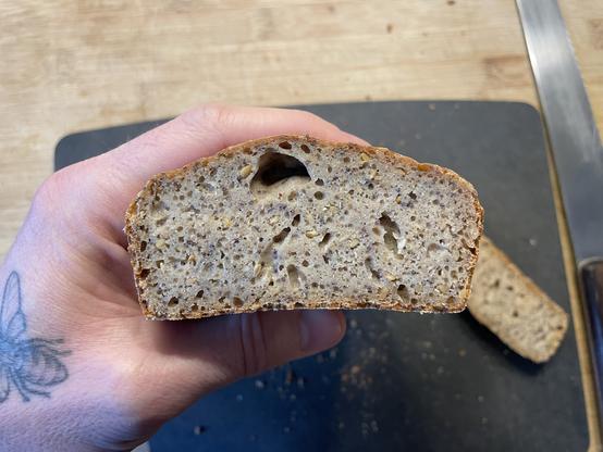 Hand holding a cut loaf of bread into the camera to show the cross cut. The bread is rectangular and small seeds are visible.