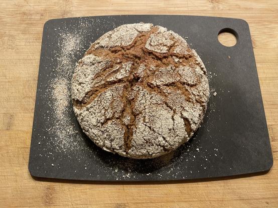 Loaf of bread shot from above. It's on a black cutting board on a wooden surface. The bread is heavily dusted with rough flour, rye meal, and shows a lot of cracks that opened up during baking. Some flour went onto the black cutting board.