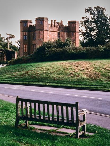 Colour portrait format photo shows a bench in the foreground, with a road crossing in front of it. Beyond a sloping grassy area the low towers of a red sandstone gatehouse (the gateway since closed up) can be seen.
