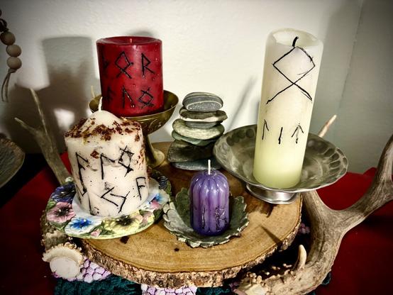 A wooden plate with two deer antlers decorated a red table. On the plate are candles with holders and trays and a stacked pile of stones.
All can have black runes carved into them. With a red, pillar candle to the right, the stones moving left and a tall white pillar candle on the far right. Below to the red candle is a shorter and wider white pillar candle with crushed petals dressing it and it sits on a pansy plated holder. To its right is a small purple candle with ridges and runes carved into it which sits on a small ashtray shaped like a flower.