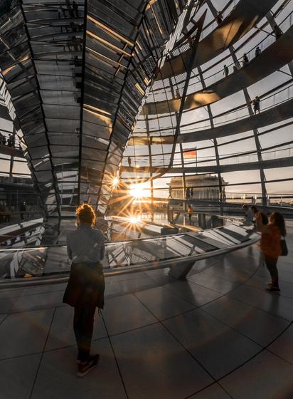 The image shows the interior of a modern building, presumably the Reichstag building in Berlin. In the center of the frame is a spiral structure made of glass and metal that twists upward. A person stands with their back to the camera, gazing at the panoramic view. The sunlight shines through the glass walls, creating a striking sunstar effect that stretches across the floor. In the background, the German flag is visible, and other people are seen walking along the upper walkway.
