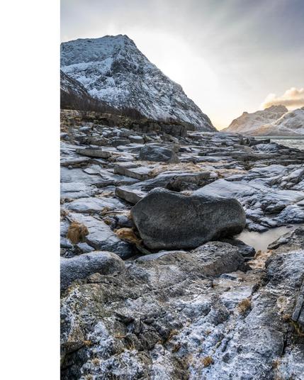 The image depicts a rugged mountainous landscape with rocky terrain in the foreground. The ground is dusted with snow, and there is a mountain peak covered in snow in the background. The sky appears to be cloudy with some sunlight illuminating the scene.