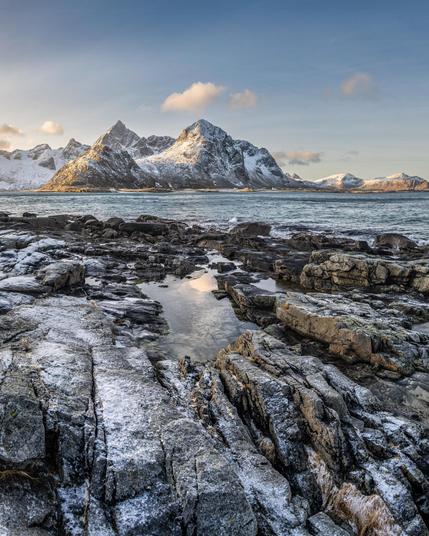 This image shows a scenic landscape featuring snow-capped mountains in the background and a rocky shoreline in the foreground. The water between the mountains and rocks reflects the sky, which has a few clouds. The lighting suggests it's either early morning or late afternoon, as the mountains are illuminated by a low sun.
